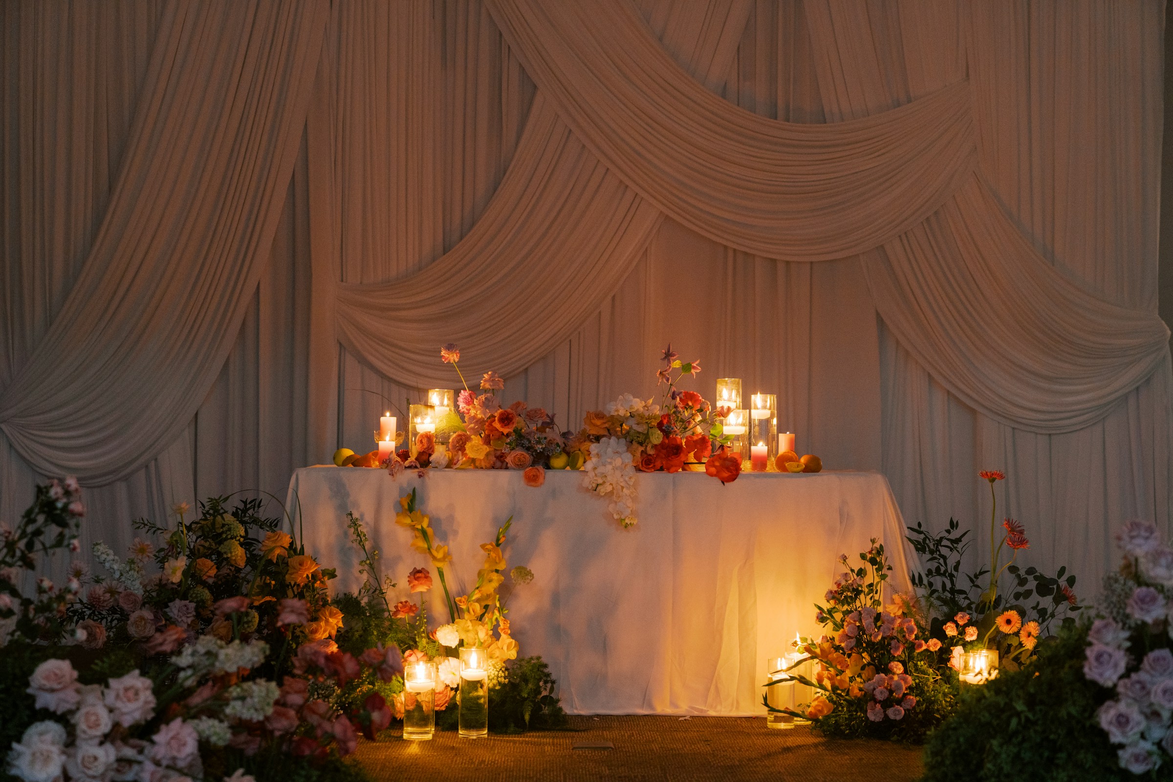 Wedding floral setup on table covered in white linen, with candles and floor covered in more flowers and white drapery