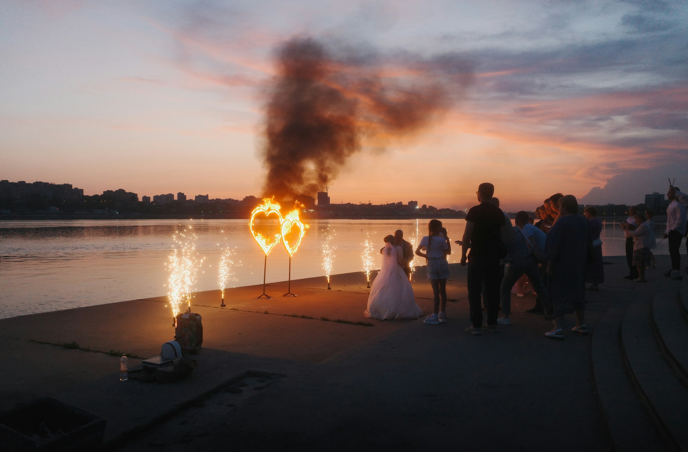 Bride and groom take photo in front heart shaped prop lit on fire, lighting up the dark outdoor evening reception